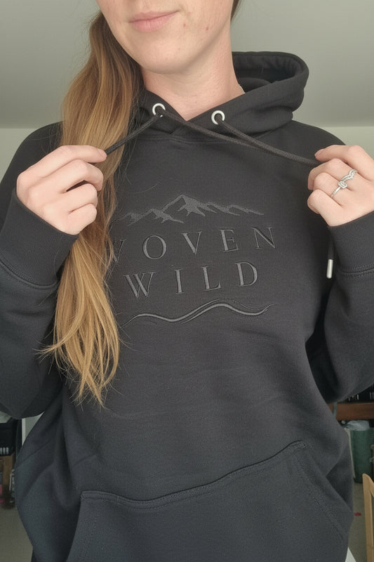 A person wearing a black hoodie with a tonal dark gray graphic on the chest that reads 'WOVEN WILD' with mountain and wave line art. The photo is taken in a casual indoor setting, showing the person's neck, hair, and hands.