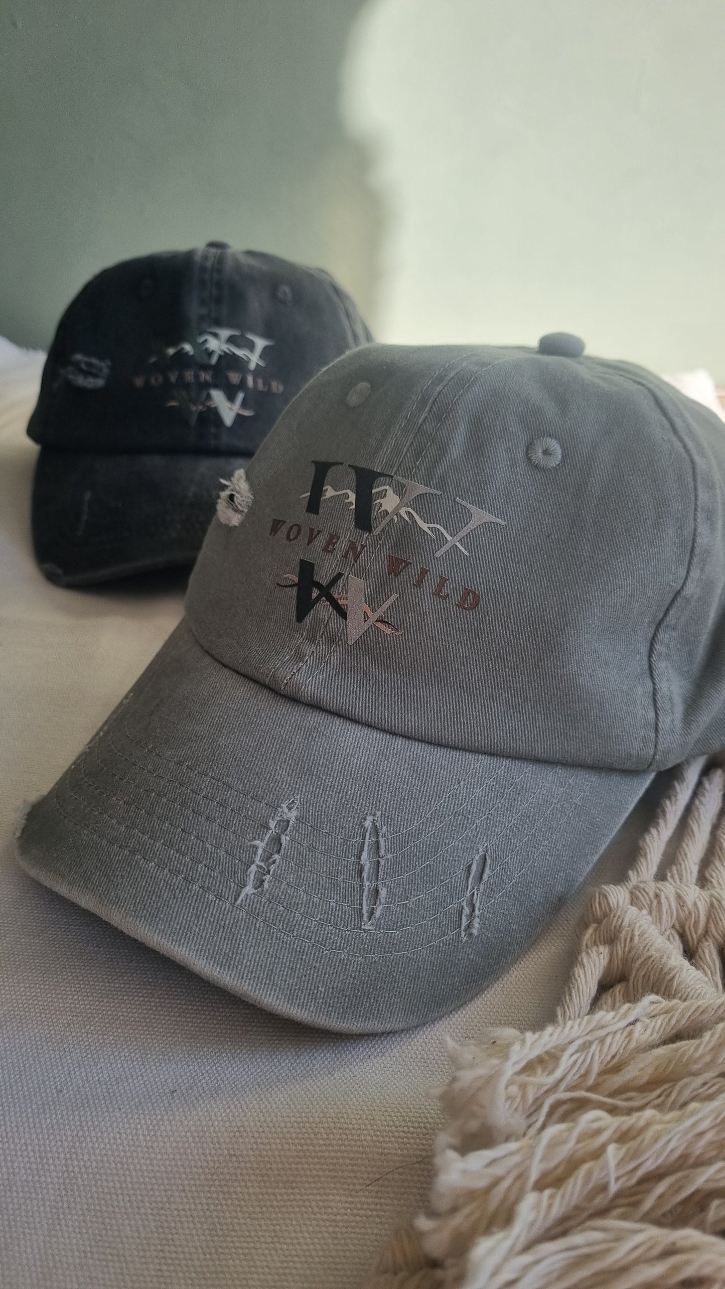 A distressed gray baseball cap with embroidered text and insignia, shown hanging against a soft beige fabric background. A second black cap is partially visible behind it.