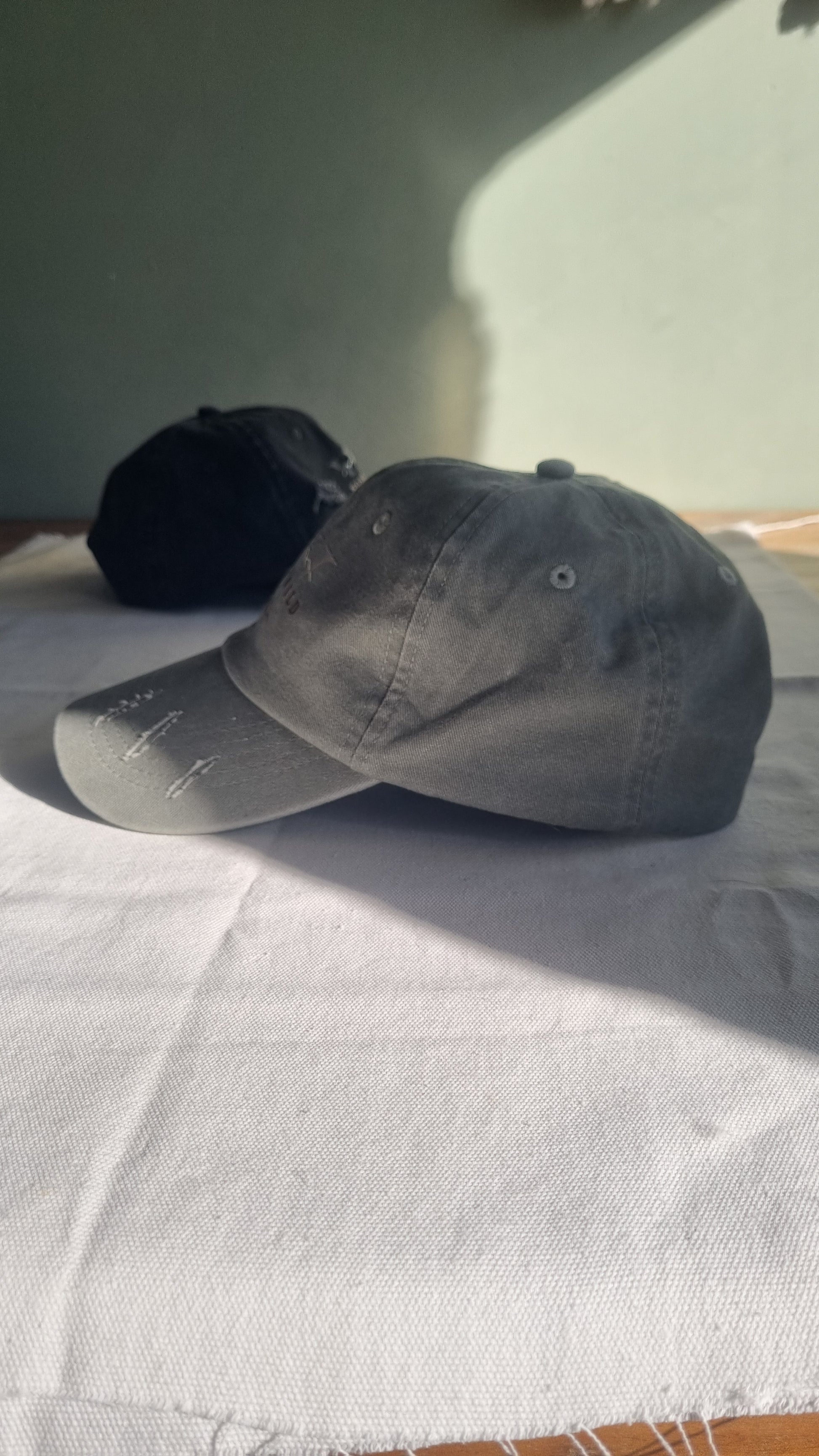 A dark gray baseball cap with a curved brim and a black cap partially tucked underneath it, resting against a white textured fabric backdrop.