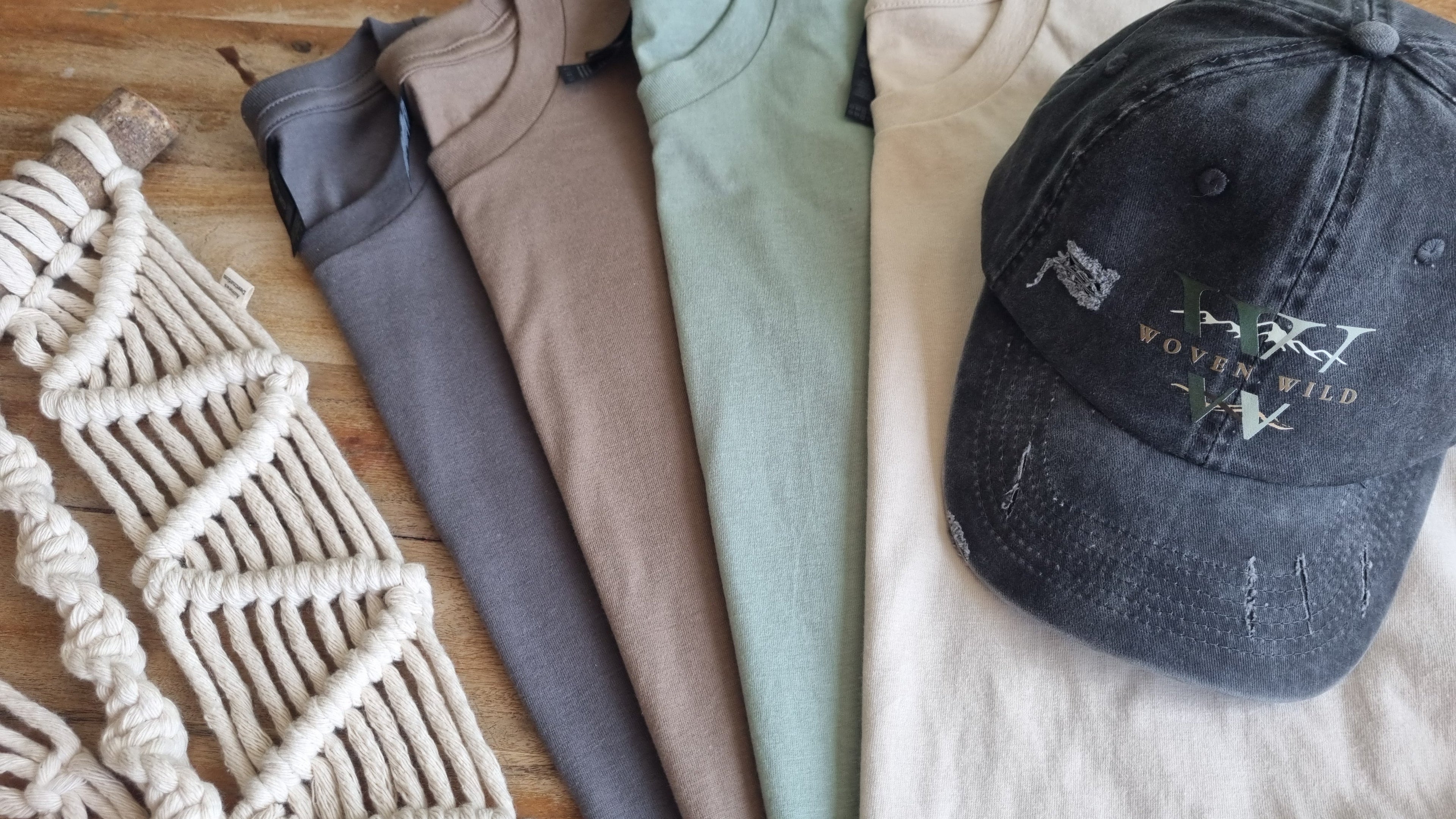 A stylish selection of organic products, including earthy tones t-shirts (washed black, coffee, turquoise and a beige 100% cotton t-shirt). Lay next to a macramé hand woven wall hanging and a washed, vintage black 'Woven Wild' cap.The Woven Wild design is nature inspired, featuring a mountain range and large monocrome letters. This photo features the sustainable, eco-friendly clothiing brand Woven Wild's products. 
