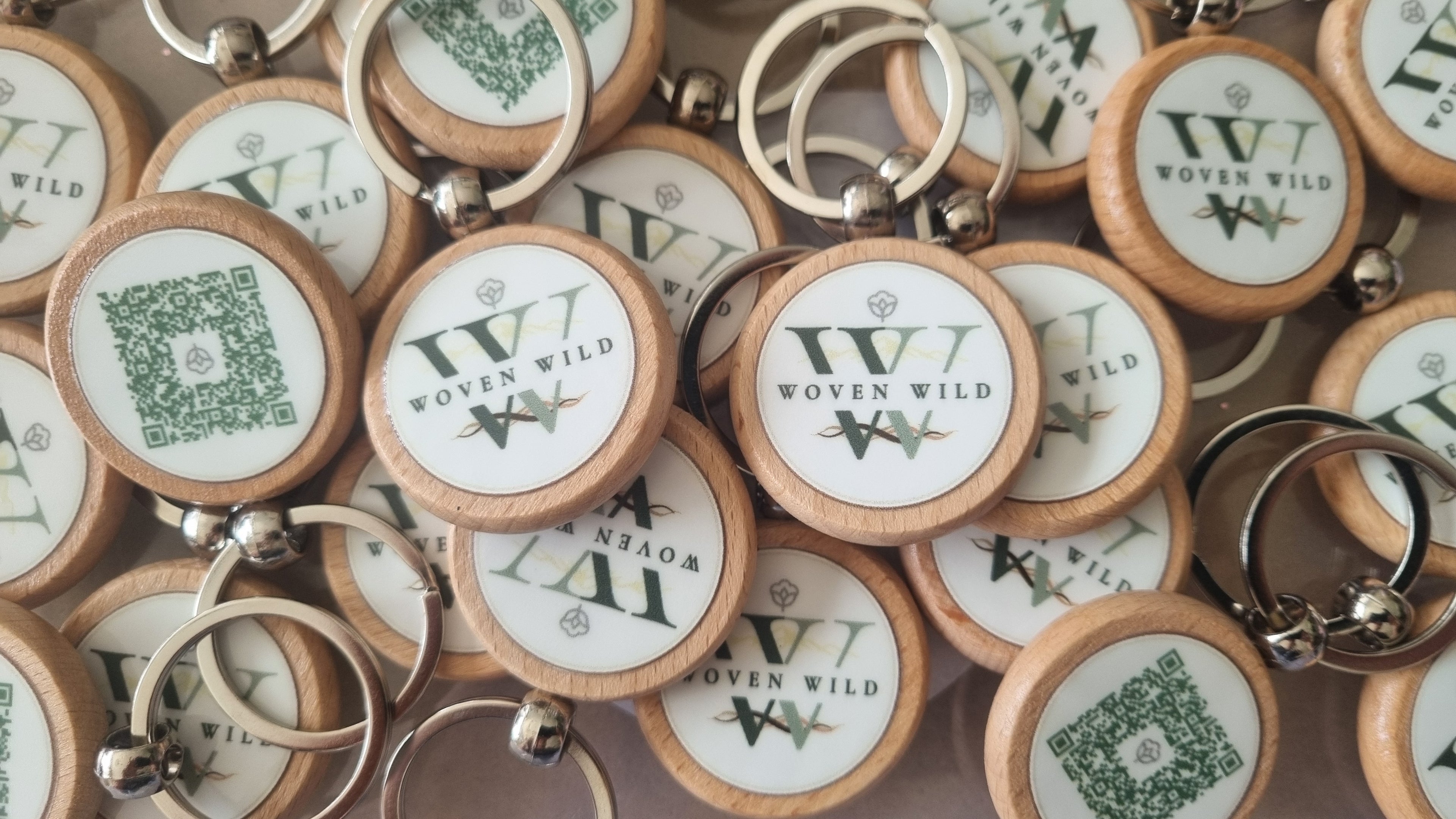 A pile of round wooden keychains with metal key rings, featuring a white center printed with the Woven Wild logo and green QR codes. This photo shows how the brand is mindful of the products they use, by using wooden products. The logo featured on these wooden key rings is the official Woven Wild logo, featuring a mountain range, selection of fluid wavey lines and monocrome letters. There is also a 100% organic cotton logo embeded in the design. 