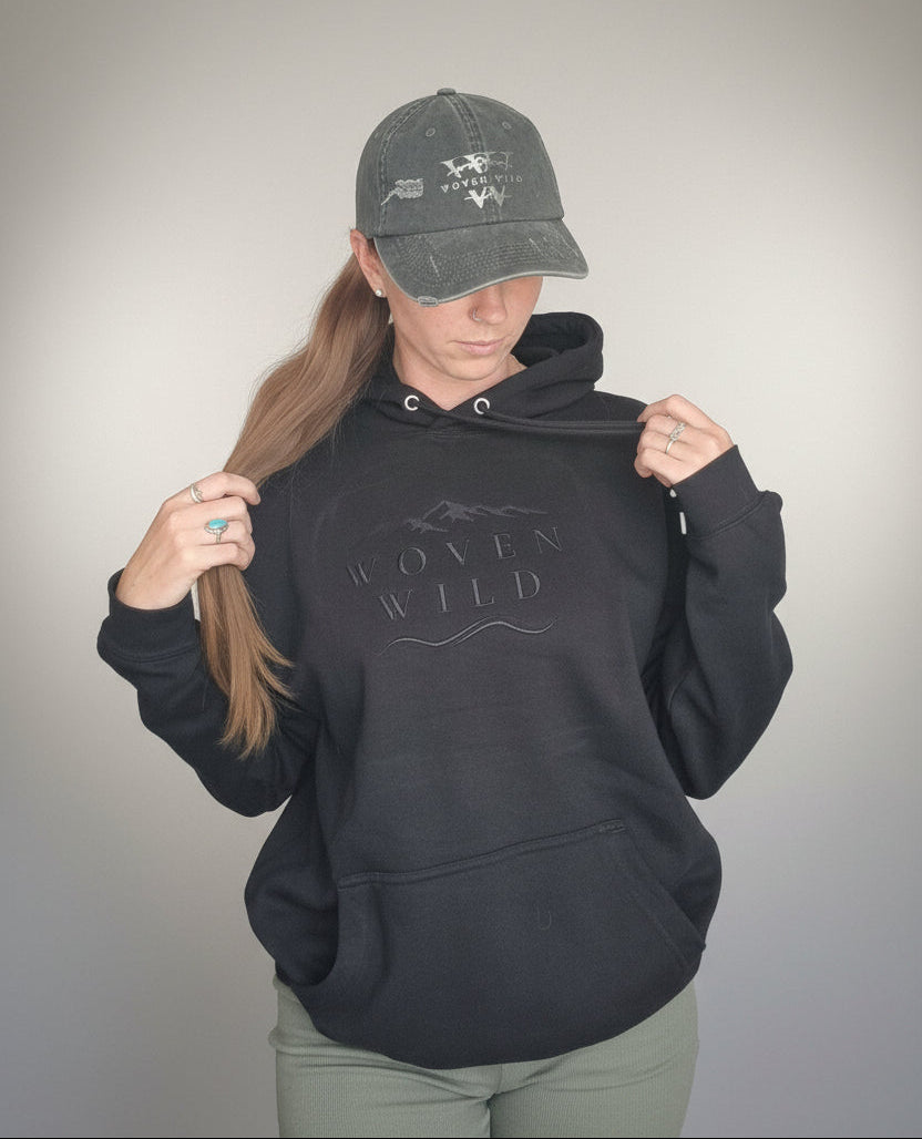 A woman wearing a black organic  pullover hoodie with a tonal 'Woven Wild' embroidered graphic on the chest, paired with a matching sage green baseball cap and olive green hiking shorts, posing against a neutral gray background.