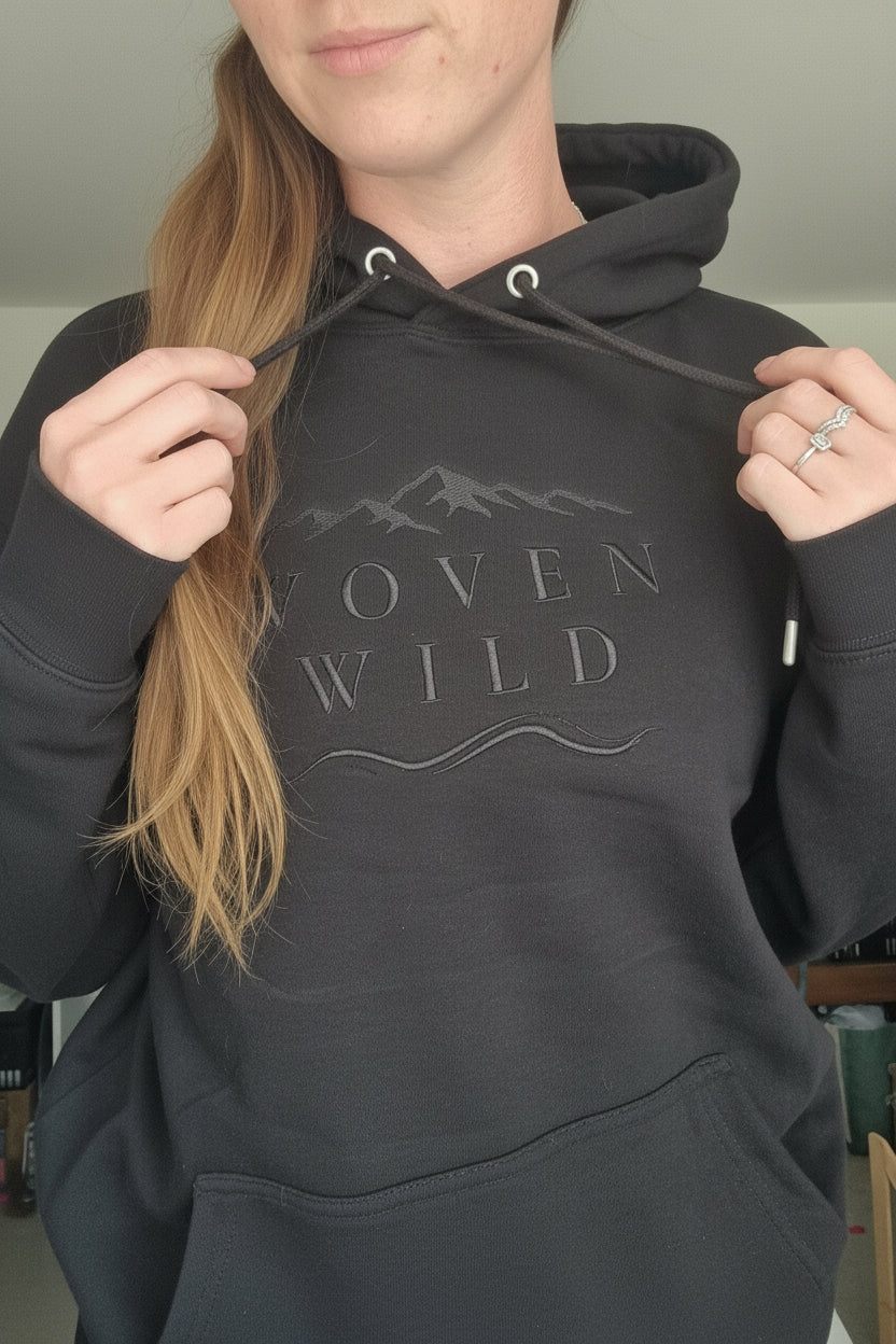 A person wearing a black hoodie with a tonal dark gray graphic on the chest that reads 'WOVEN WILD' with mountain and wave line art. The photo is taken in a casual indoor setting, showing the person's neck, hair, and hands.
